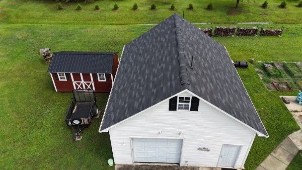 Aerial view of a dark gray architectural shingle roof on a white garage with a barn and utility trailer nearby.
