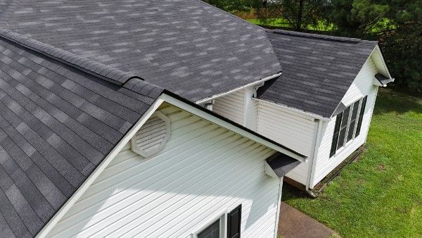 Aerial view of a gray asphalt shingle roof with white vinyl siding, a round gable vent, and downspouts.