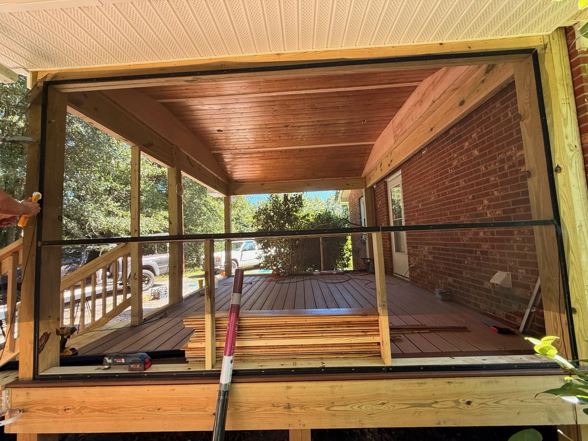 A wood framed screened porch with dark composite decking, a wood plank ceiling, and stacked lumber against a brick house.