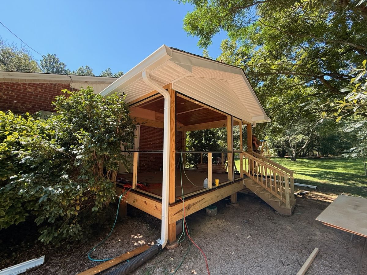A wood framed covered porch addition with a shingled gable roof, vinyl soffit, and gutters attached to a brick house.