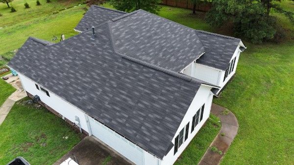 Aerial view of a dark gray asphalt shingle roof on a white house surrounded by green lawn and trees.