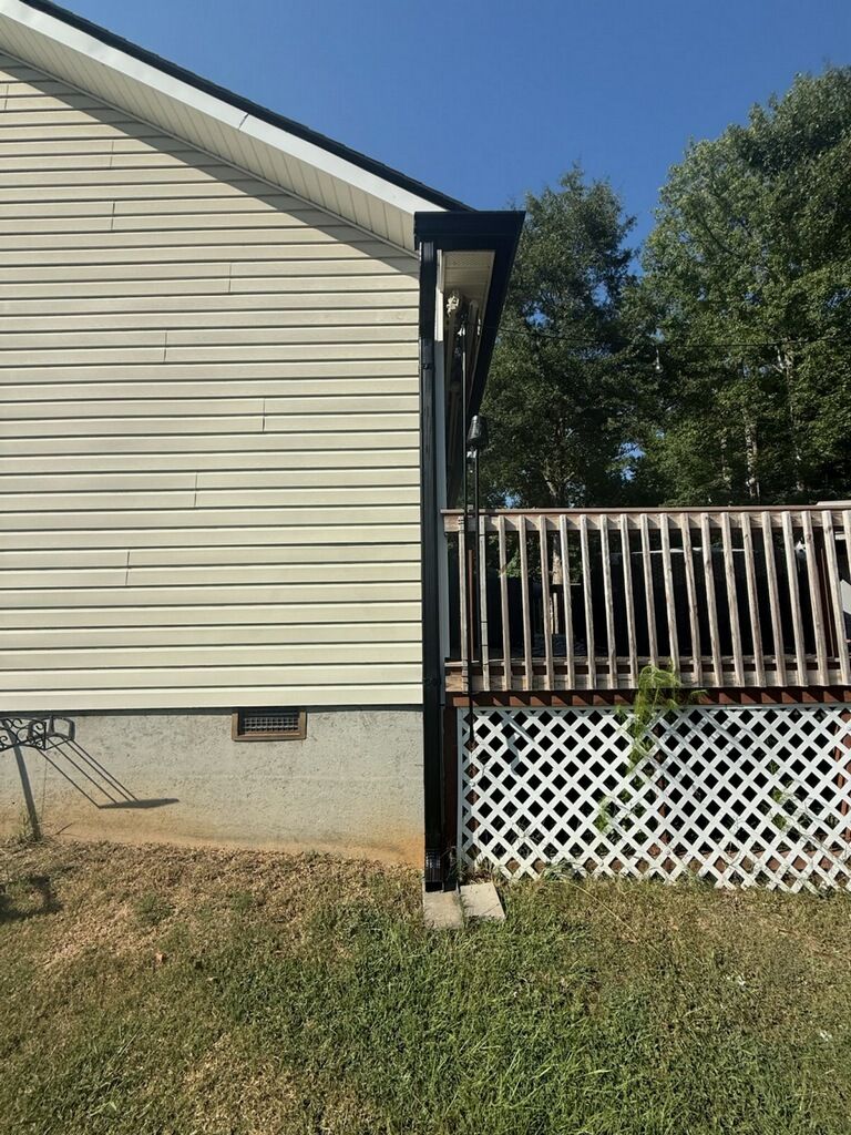 A black downspout runs down the corner of a beige vinyl-sided house next to a wooden deck with white lattice skirting.