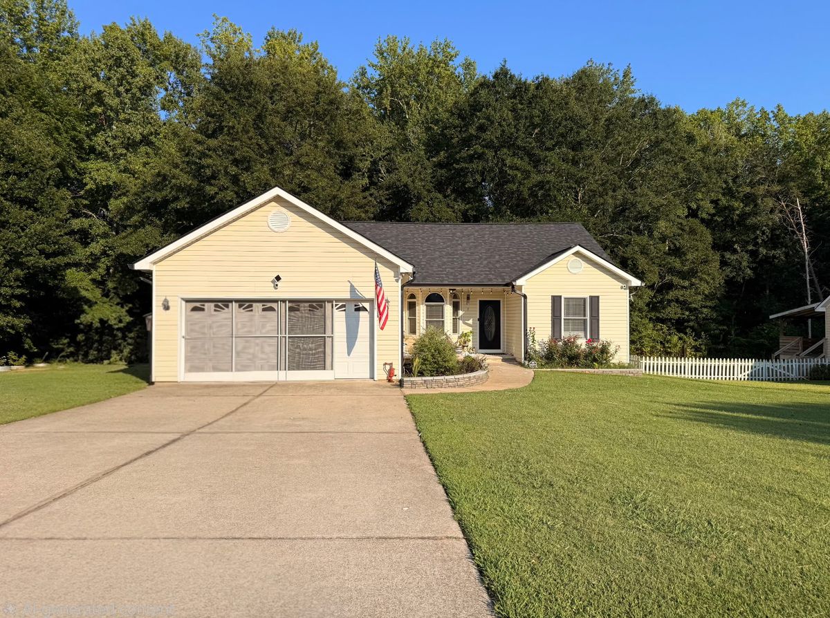 Single story yellow vinyl siding home with dark shingle roof, attached garage, and white picket fence along the yard.